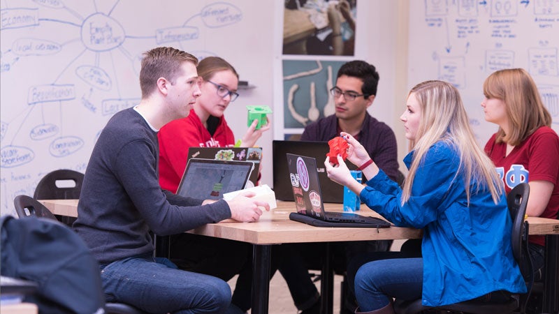 Five students around a table, working on a group project.  White board has lots of notes.